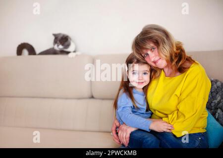 Sorridi la nonna di mezza età e la nonna piccola che abbraccia e guarda la macchina fotografica mentre si siede sul divano con il gatto carino sullo sfondo mentre Foto Stock