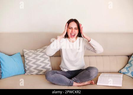 Infastidita giovane donna seduta sul divano e gridando mentre lavora a distanza con il laptop e i documenti a casa durante l'isolamento Foto Stock