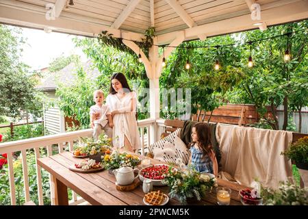 Donna sorridente seduta su panchina con adorabili fratelli al tavolo di legno durante la colazione in cortile Foto Stock