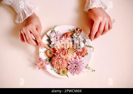 Vista dall'alto di una femmina anonima con taglio a forchetta e a coltello e mangiare fiori naturali su piatto contro sfondo di pesca Foto Stock