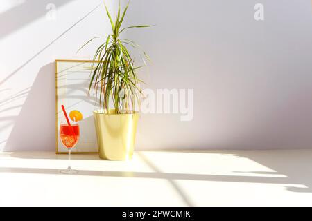 Bicchiere di cocktail di frutta fresca collocato al piano vicino alla pianta in vaso e cornice vuota contro la parete bianca nella stanza illuminata dal sole Foto Stock