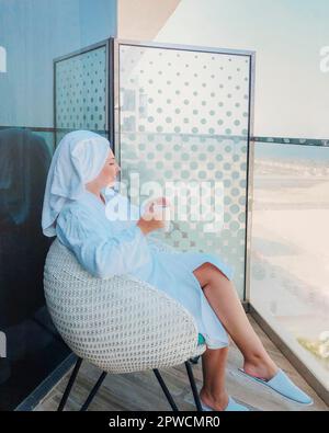 Vista laterale della giovane donna in accappatoio bianco e asciugamano sulla testa seduta su poltrona con tazza di caffè sul balcone Foto Stock