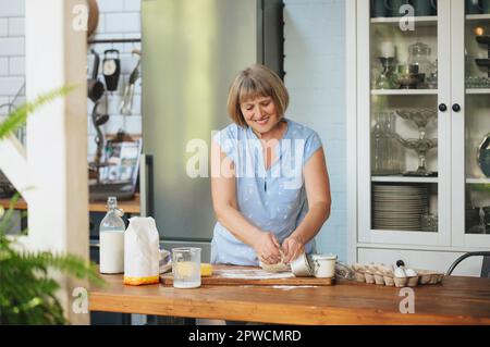 Casalinga donna anziana felice che fa impastare pasta per torta mentre si trova dietro un tavolo di legno in cucina con ingredienti per la cottura, sorridendo Foto Stock