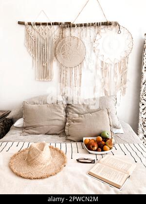 Interni moderni in stile scandi boho con cuscini decorativi, cappello in vimini, frutta e libro aperto sul letto, decorazioni in macrame e dreamcatcher Foto Stock