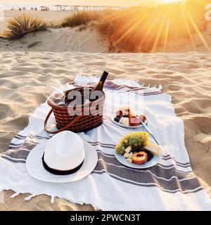 Romantico pic-nic sulla spiaggia sabbiosa al tramonto della sera, coperta con bottiglia di vino e bicchieri in cesto di vimini, cibo, frutta e spuntini sui piatti Foto Stock