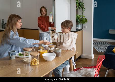 Una bambina adora che tiene una bottiglia di latte dal frigorifero per una colazione in famiglia con mamma e fratello Foto Stock