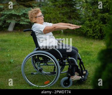 Donna caucasica anziana che fa esercizi mentre si siede su una sedia a rotelle all'aperto. Foto Stock