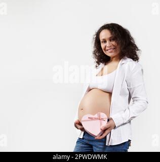 Donna incinta sorridente dai capelli ricci che tiene la scatola del regalo a forma di cuore rosa vicino al suo ventre, isolata su fondo bianco Foto Stock