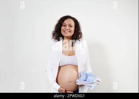 Donna incinta che tocca delicatamente la pancia, sorridendo alla macchina fotografica, posando con stivaletti blu a maglia su sfondo bianco. Foto Stock