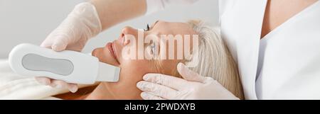 Il cosmetologo professionale sta facendo la pulizia ultrasonica del viso per bella donna matura caucasica felice in clinica Foto Stock
