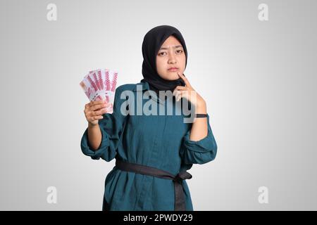 Ritratto di confuso asiatico hijab donna in vestito casual mostrando centomila rupiah mentre pensando con mano sul mento. Finanziario e risparmio co Foto Stock