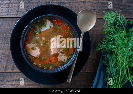 Zuppa di salmone fresco con carote, patate e peperoni rossi in un piatto nero su tavolo di legno, primo piano. La cena gustosa consiste in una zuppa di pesce con sa Foto Stock