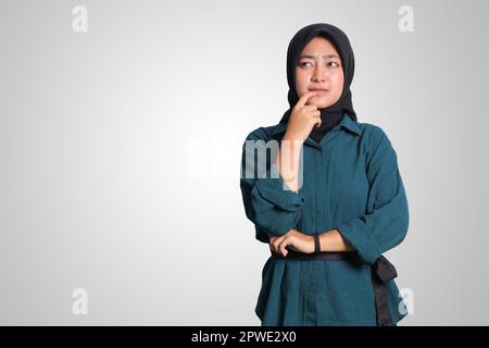 Ritratto di donna musulmana asiatica confusa con hijab in piedi contro sfondo bianco, guardando in alto e pensando alla domanda con mano sul mento Foto Stock