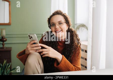 Donna felice che usa la tecnologia del telefono cellulare a casa, sorridente mentre controlla le app sul suo smartphone e si rilassa con i dispositivi cellulari in mano. Foto Stock