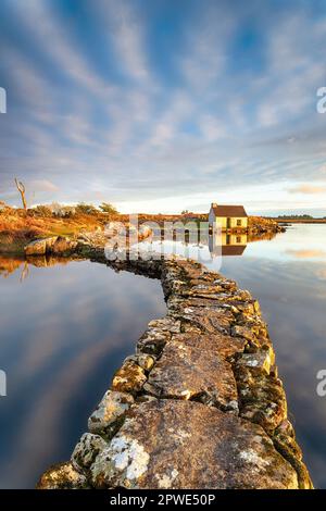 Un vecchio molo di pietra attraverso un lago che conduce ad un piccolo cottage nel Parco Nazionale di Connemara in Irlanda Foto Stock