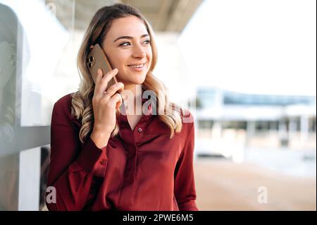 Chiamata telefonica. Splendida donna caucasica elegante, donna di successo, si trova all'aperto, utilizza il suo smartphone, parlando con un amico, collega o cliente, guarda via, sorridente amichevole Foto Stock