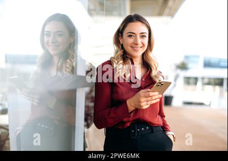 Splendida ed elegante donna caucasica sta all'aperto, utilizza il suo smartphone, scrive un messaggio, invia messaggi con gli amici nei social media, cerca informazioni, guarda la fotocamera, sorridendo amichevole Foto Stock