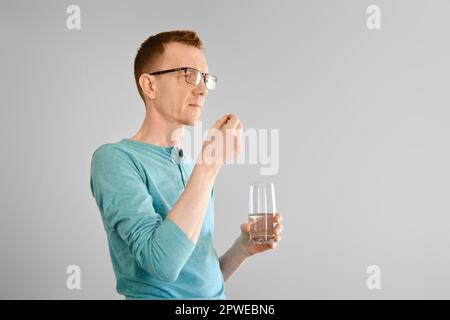 Uomo di mezza età in piedi con un bicchiere d'acqua e prende la pillola dal mal di testa Foto Stock