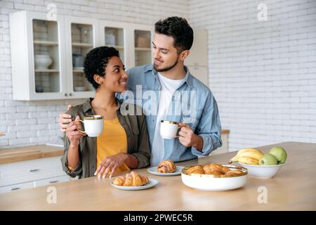 Felice marito e moglie di diverse nazionalità hanno gustoso fine settimana colazione in cucina a casa con croissant e caffè, godendo di trascorrere il tempo insieme, si guarda con amore, sorriso Foto Stock