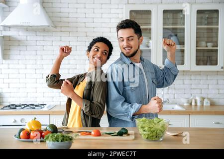 Felice coppia innamorata è cucinare a casa in cucina insieme. L'uomo caucasico e la donna afroamericana si divertono, ballano mentre fanno insalata fresca e sana per un pranzo, godendosi il fine settimana Foto Stock