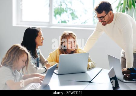 Educatore informatico che guida un gruppo di giovani studenti in una classe di codifica. Gli insegnanti si impegnano con i bambini mentre imparano a scrivere codice sui loro laptop, Foto Stock