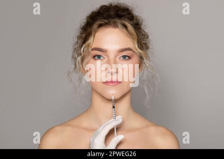 Bella donna caucasica che ottiene l'iniezione facciale, guardando la macchina fotografica mentre il cosmetologist tiene la siringa vicino al mento Foto Stock