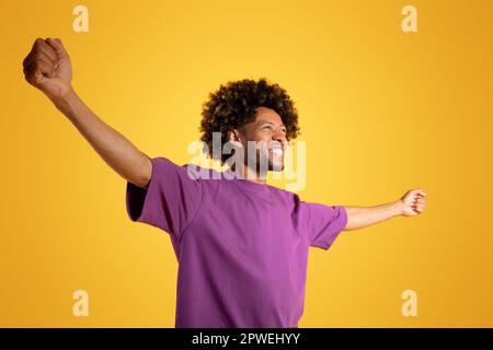 Sorridente eccitato adulto afroamericano riccio uomo in viola t-shirt solleva le braccia ai lati gode di libertà Foto Stock