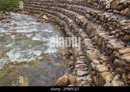 Rafforzare le rive del fiume di montagna, rete e pietre. Riva del fiume paesaggio e grandi pietre con rete metallica Foto Stock