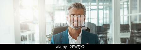 Uomo d'affari maturo sorridente che mostra le dita sollevate mentre si siede in ufficio Foto Stock