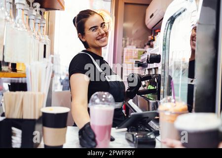 Giovane barista sorridente in bicchieri, abiti casual, grembiule e guanti neri che consegnano un cocktail matcha rosa alla sua cliente. Bella ragazza che lavora in s Foto Stock