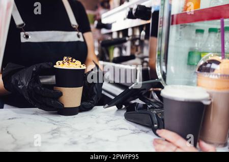Primo piano le mani del barista femminile in guanti neri. La donna in grembiule sta consegnando al suo cliente un cocktail di caffè preparato con crema e patatine di cioccolato. Scheda Foto Stock