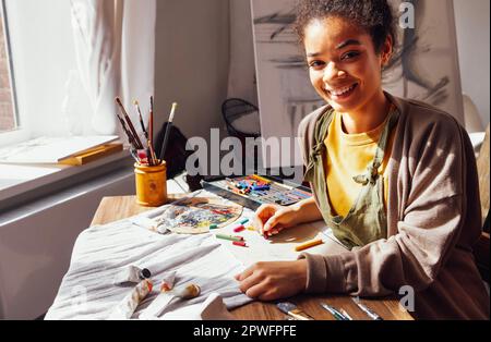 Giovane artista ragazza afroamericana creativa dipinge con colori pastello mentre si siede al tavolo di fronte alla finestra. Bella pittore femminile in casual w Foto Stock