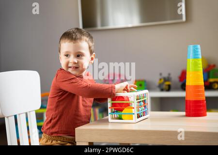 Un ragazzino carino di due anni gioca con palle multicolore e si siede al tavolo di un bambino in camera. Sfere sensoriali tattili. Istruzione Foto Stock