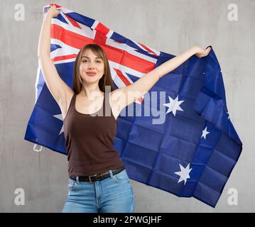 Felice giovane donna allegra che sventola bandiera nazionale dell'Australia mentre guarda la macchina fotografica sullo sfondo grigio della parete Foto Stock