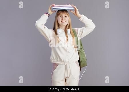 Ritratto di ragazza felice scuola trendy in tuta beige con zaino, libri di lavoro e cuffie isolato sul grigio. Foto Stock