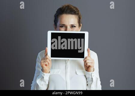 Donna trendy di mezza età in blusa bianca con schermo vuoto del tablet PC isolato su sfondo grigio. Foto Stock