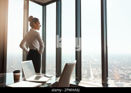 Ceneri in cima al mondo. Ripresa retroversa di una giovane e attraente donna d'affari in piedi con le mani sui fianchi in ufficio. Foto Stock