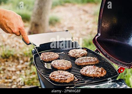 Primo piano di un maschio che flipping spesso hamburger di manzo frittura sulla griglia all'aperto, barbecue nel parco Foto Stock