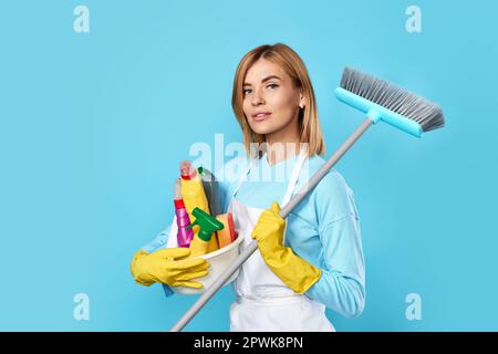 donna bionda in guanti di gomma gialla e grembiule detergente che tiene secchio di detergenti e scopa su sfondo blu. Foto Stock