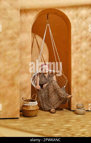 Vecchia donna attiva con capelli rosa siede in vimini swing in arco Foto Stock
