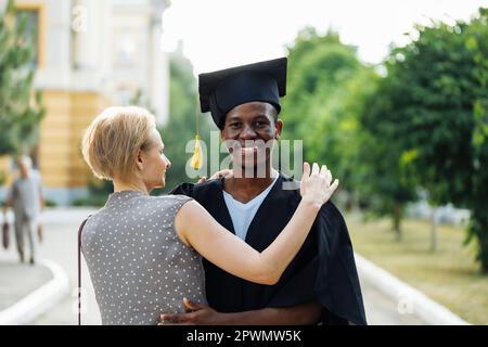 Gioioso afro studente americano in mantello di laurea e cappello gratefully abbracciando la donna di mezza età. Istruzione superiore, laureato all'università, pensieri Foto Stock