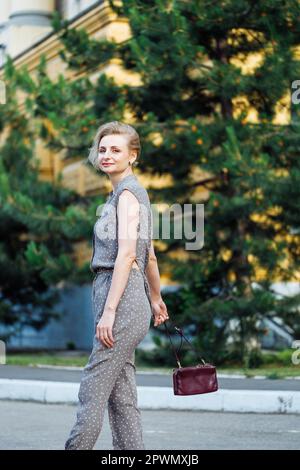 Ritratto vista laterale di una donna di 40 anni che cammina all'aperto tra abeti verdi con borsa a spalla in pelle di borgogna in mano. Signora di mezza età in bella Foto Stock