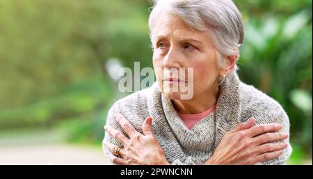 È un po' freddo oggi. una donna anziana spensierata seduta su una panchina mentre contempla fuori in un parco Foto Stock