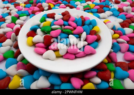 Un piatto bianco con caramelle multicolore a forma di cuore (primo piano), sullo sfondo delle stesse caramelle multicolore. Vista dall'alto Foto Stock