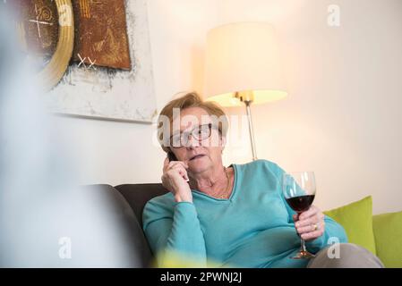 Vecchia donna che si rilassa sul divano con un bicchiere di vino rosso e parla al telefono Foto Stock