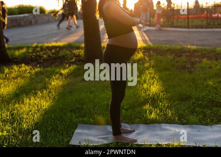 Vista laterale di giovane donna che aspetta il bambino in abito nero si leva sul tappeto scalda e medita in caldo sole giorno d'estate. Concetto di sport Foto Stock