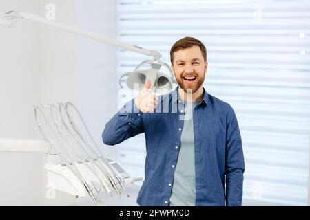 Ritratto di paziente felice vicino a sedia dentale e mostra un gesto con un pollice in classe, buono. Odontoiatria moderna con l'uso di nuove tecnologie. Foto Stock