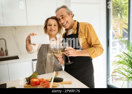 Coppia adulta senior che prende selfie su smartphone mentre cucinano e bevono vino in cucina Foto Stock