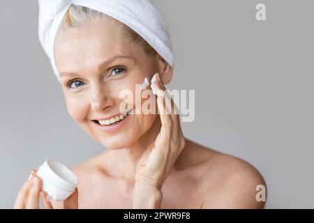 Bella donna matura applicando crema idratante sotto i suoi occhi, facendo il trattamento di bellezza con il prodotto cosmetico Foto Stock