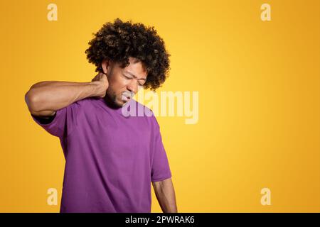 Triste uomo afroamericano riccio in t-shirt viola preme la mano al collo e soffre di dolore muscolare Foto Stock